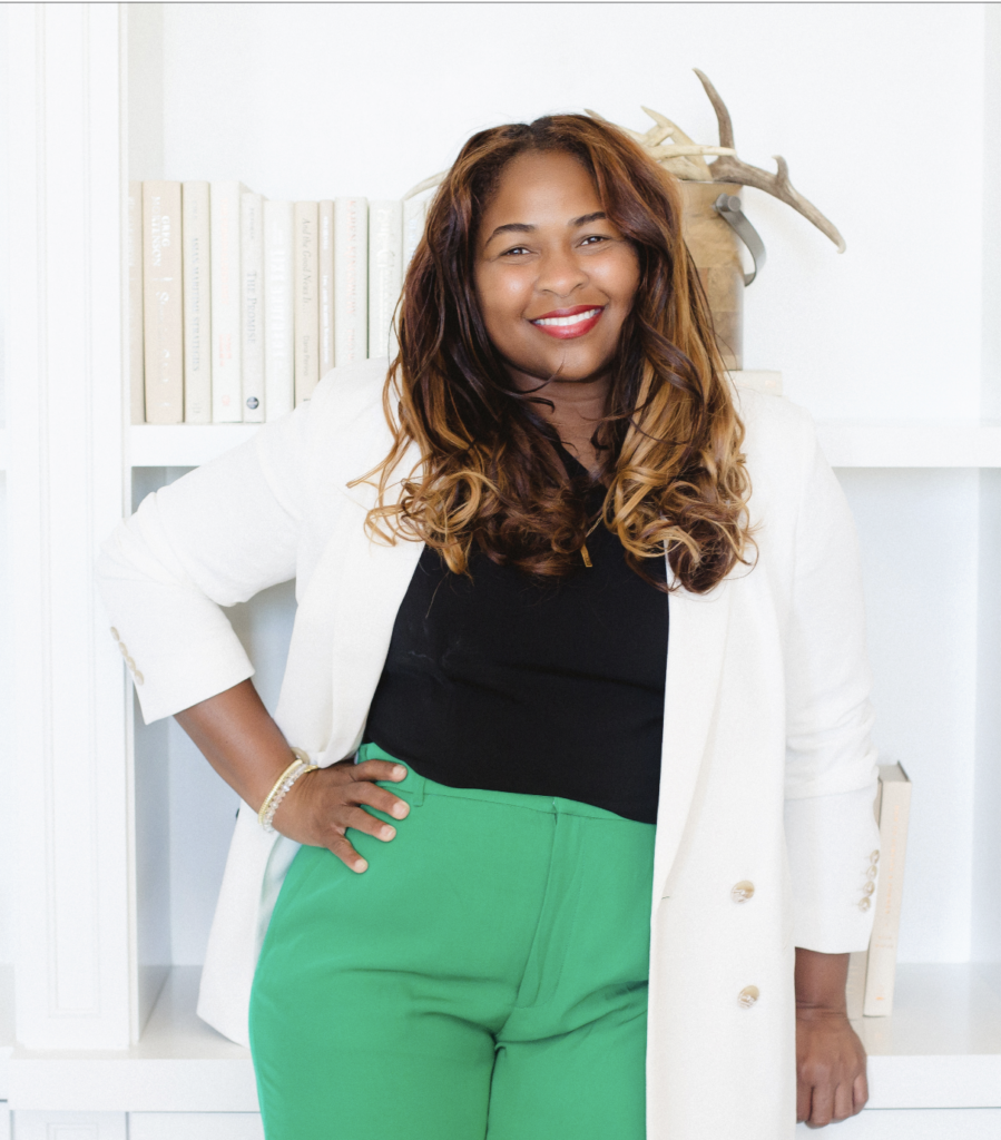 Beautiful black woman with long hair smiling with her hand on her hip. Wearing a white blazers, black top and green pants.