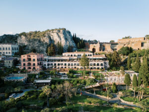 A stunning aerial view of Grand Hotel Timeo in Taormina, Italy, with its lush gardens, multiple terraces, and the backdrop of ancient ruins and rocky hills.