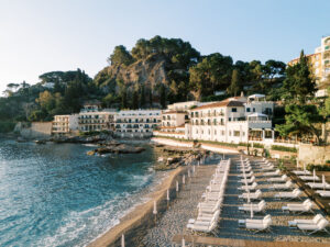 A scenic view of Villa Sant'Andrea, a luxurious hotel on the Sicilian coast, featuring white buildings, lush greenery, and a private beach with rows of lounge chairs.