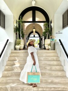 Michelle Norwood in a white maxi dress and straw hat ascends a staircase at Belmond Cap Juluca in Anguilla, holding a teal and beige tote bag.