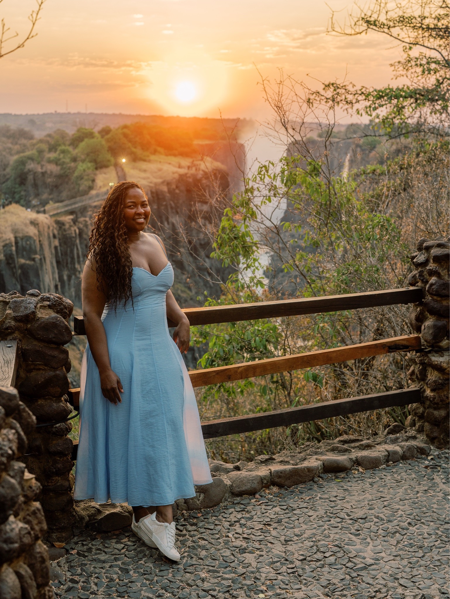 Traveler standing at a scenic overlook during sunset, wearing a light blue dress with white sneakers, surrounded by nature.