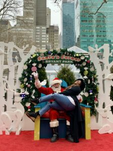 Michelle Norwood joyfully sitting on Santa's lap at Santa's Corner in Bryant Park, surrounded by festive holiday decorations and a wreath with skyscrapers in the background.